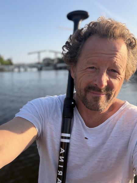 A man with curly hair and a beard smiles at the camera, holding a paddle, against the backdrop of the Amstel river in Centrum, Amsterdam